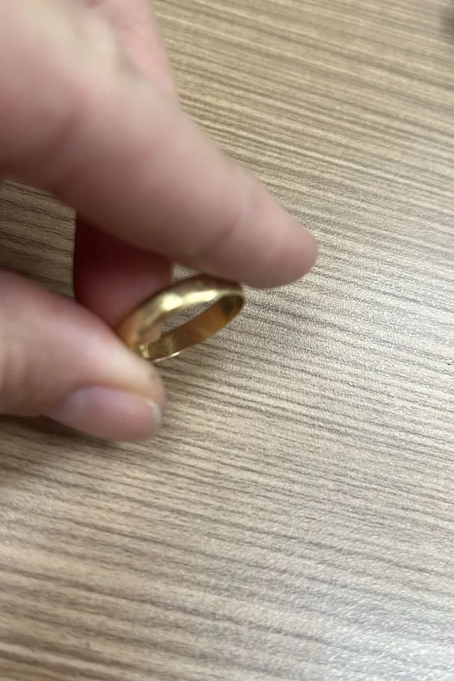 Fotografia de referência da aliança procurada, segurada entre os dedos sobre uma mesa de madeira